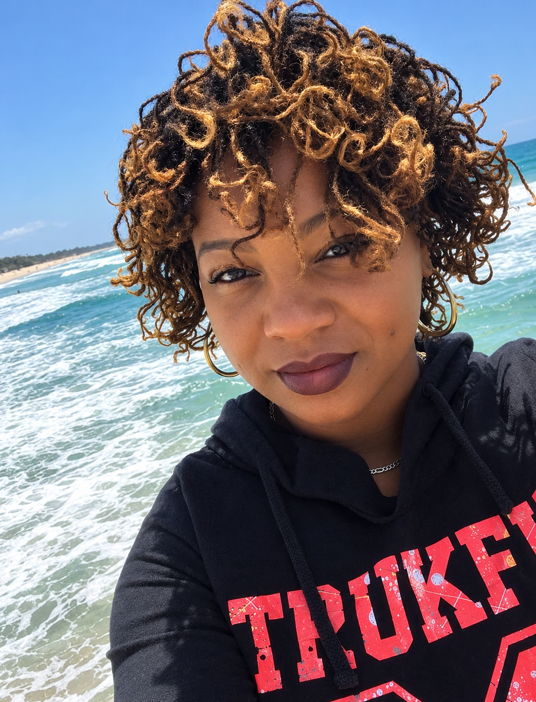 Client with curly, honey-highlighted Sister Locs photographed in front of the ocean